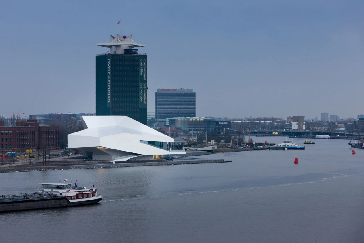 White angular riverside building with a tall dark tower behind, city skyline in distance; boat on calm river.