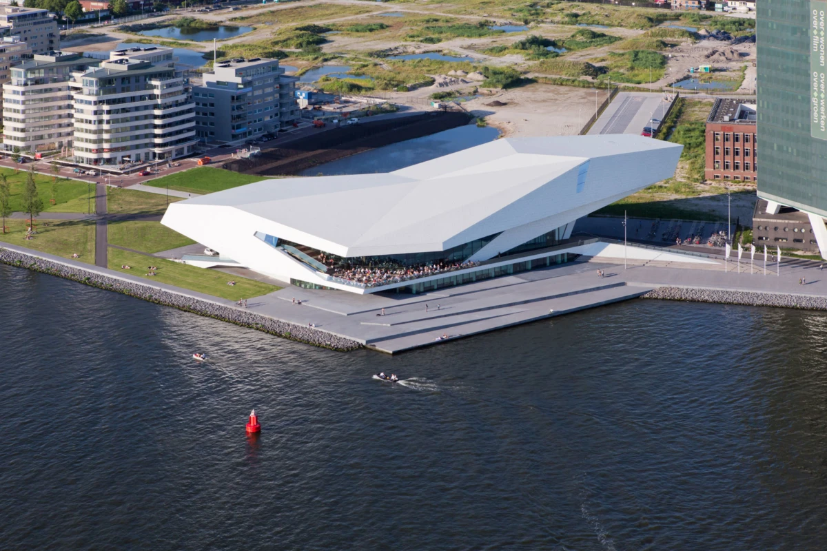 Modern angular white riverside concert hall with terrace spectators, by a harbor with boats.