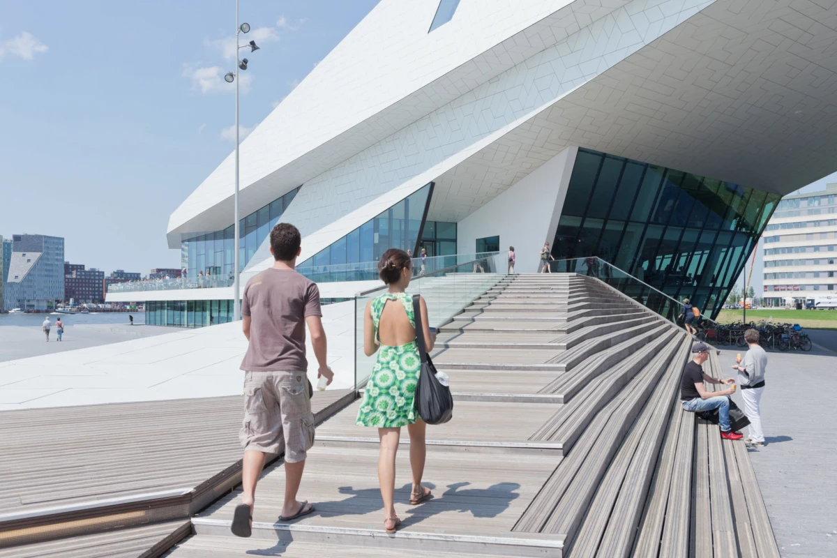 Man and woman walk up wide wooden steps toward a futuristic white building by the waterfront.