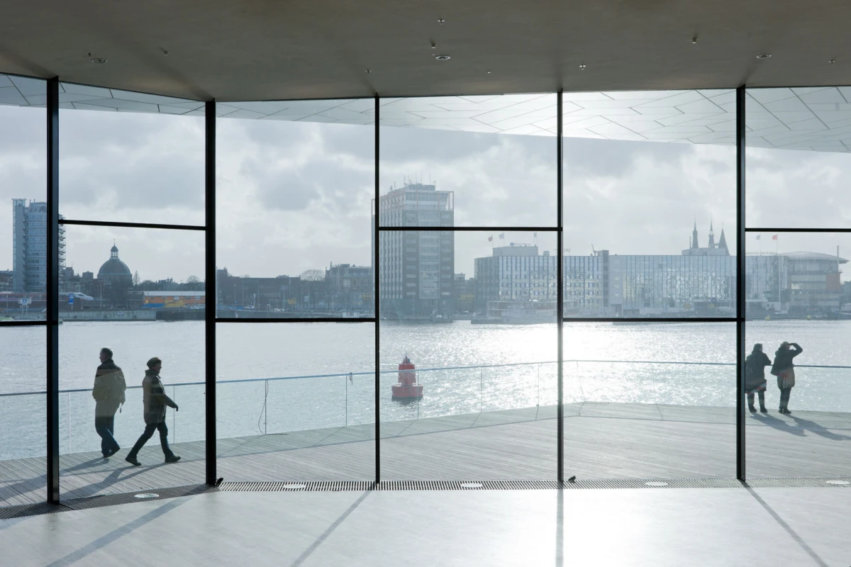 Two pedestrians walk along a waterfront promenade seen through floor-to-ceiling glass walls; city skyline across the water; red buoy.