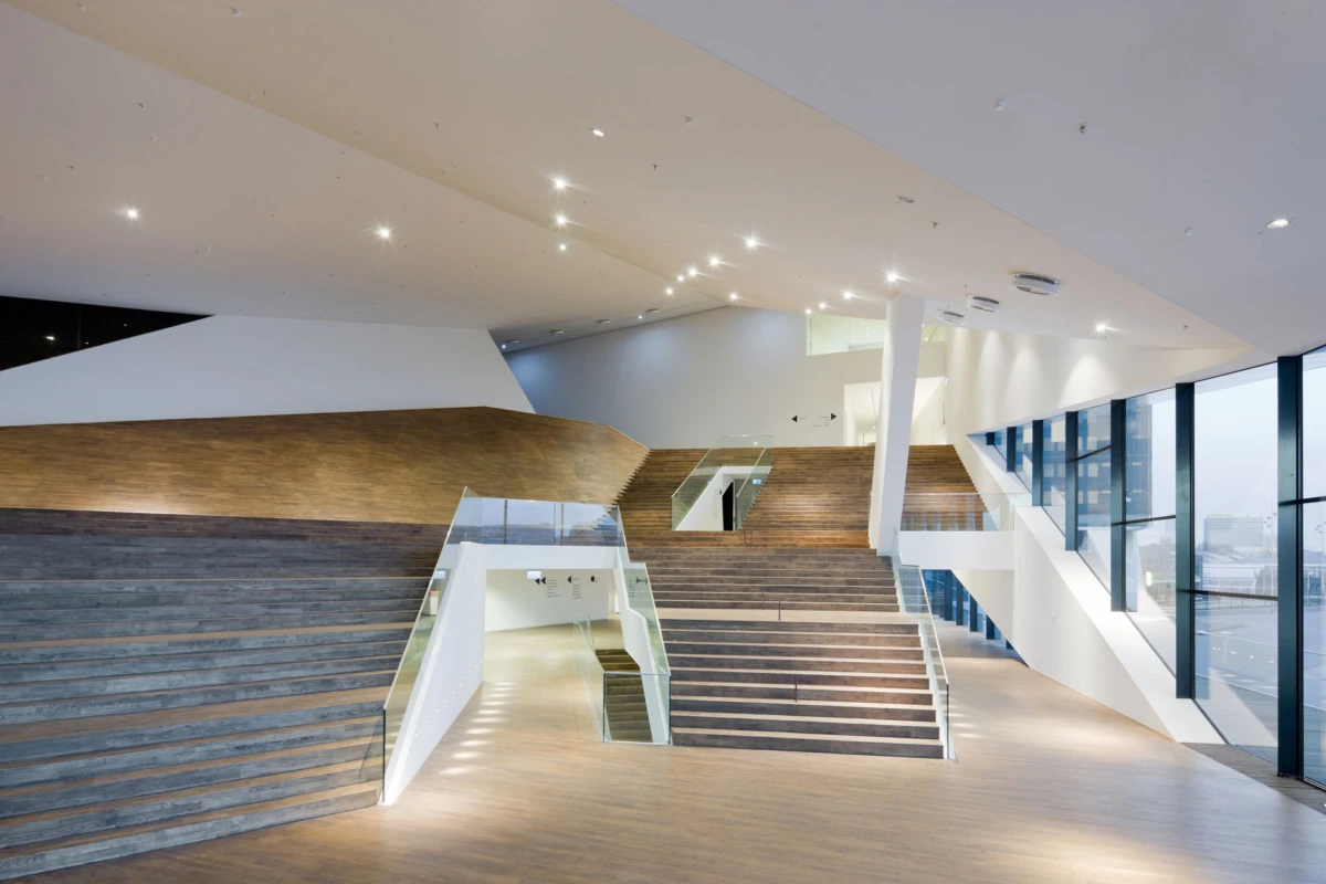 Empty modern atrium with a grand wooden staircase, glass railings, and expansive windows.