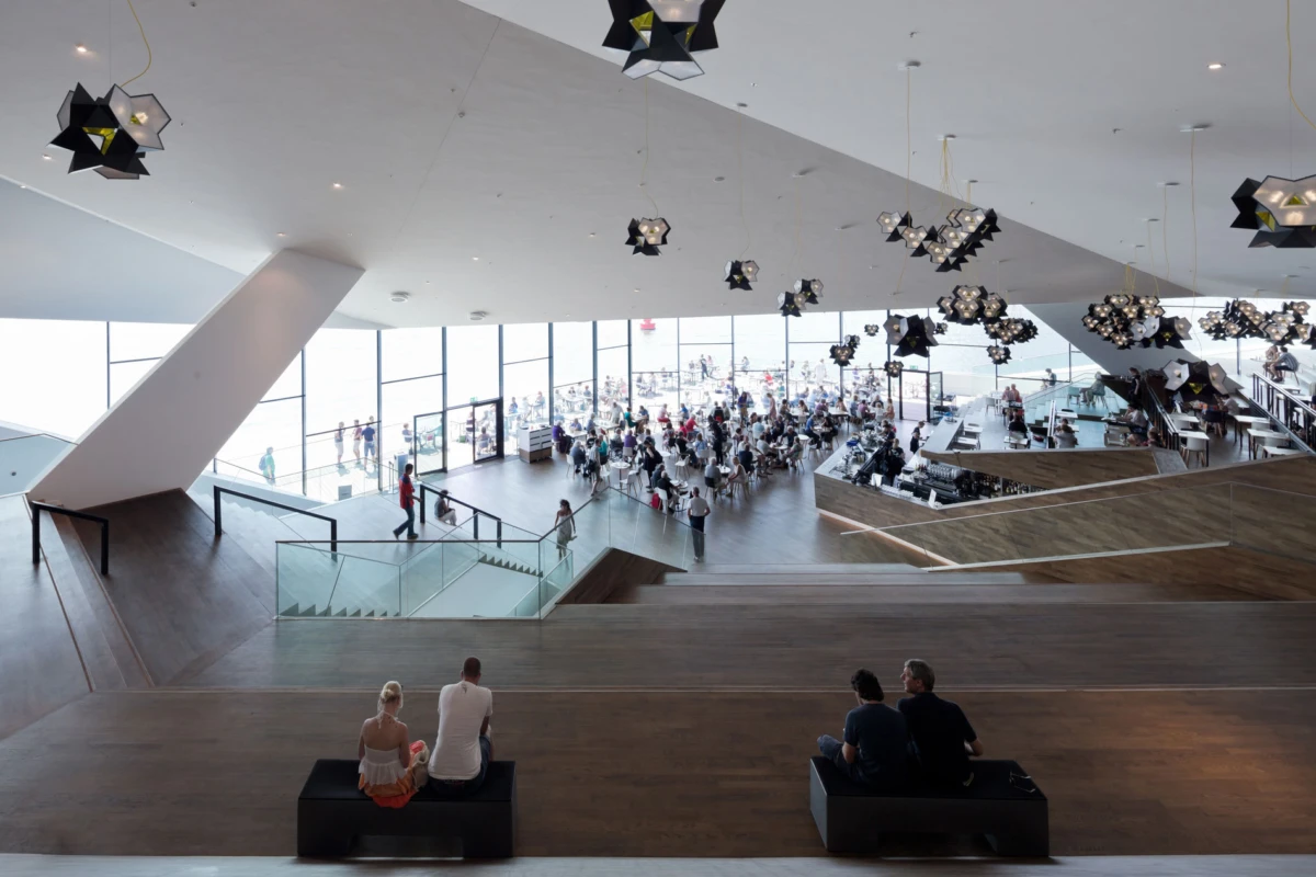 Two couples sit on benches in the foreground of a large modern atrium with wooden floors, glass railings, and a busy seating area under geometric pendant lights.