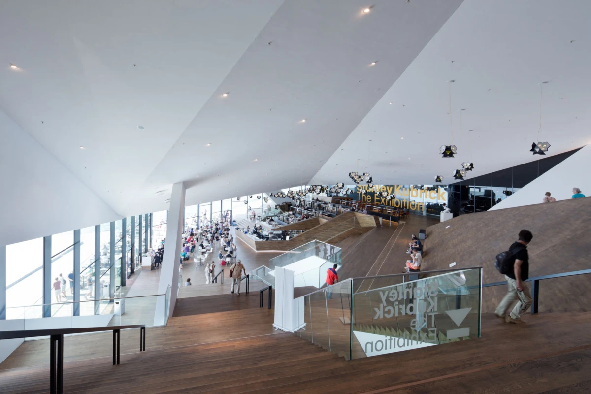 Modern museum interior with visitors walking on wooden ramps; prominent sign reads Stanley Kubrick: The Exhibition.