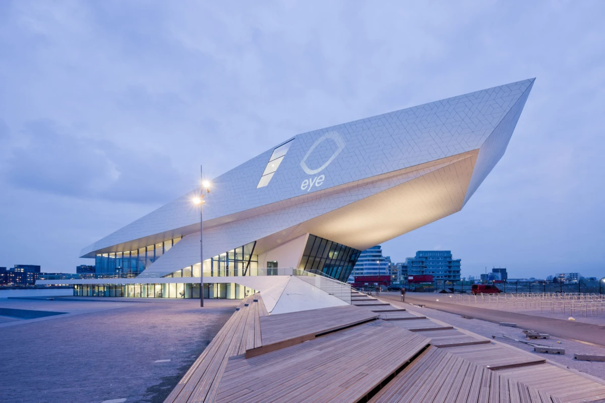 Futuristic pavilion with a slanted roof and glass walls on a wooden boardwalk along a waterfront at dusk.