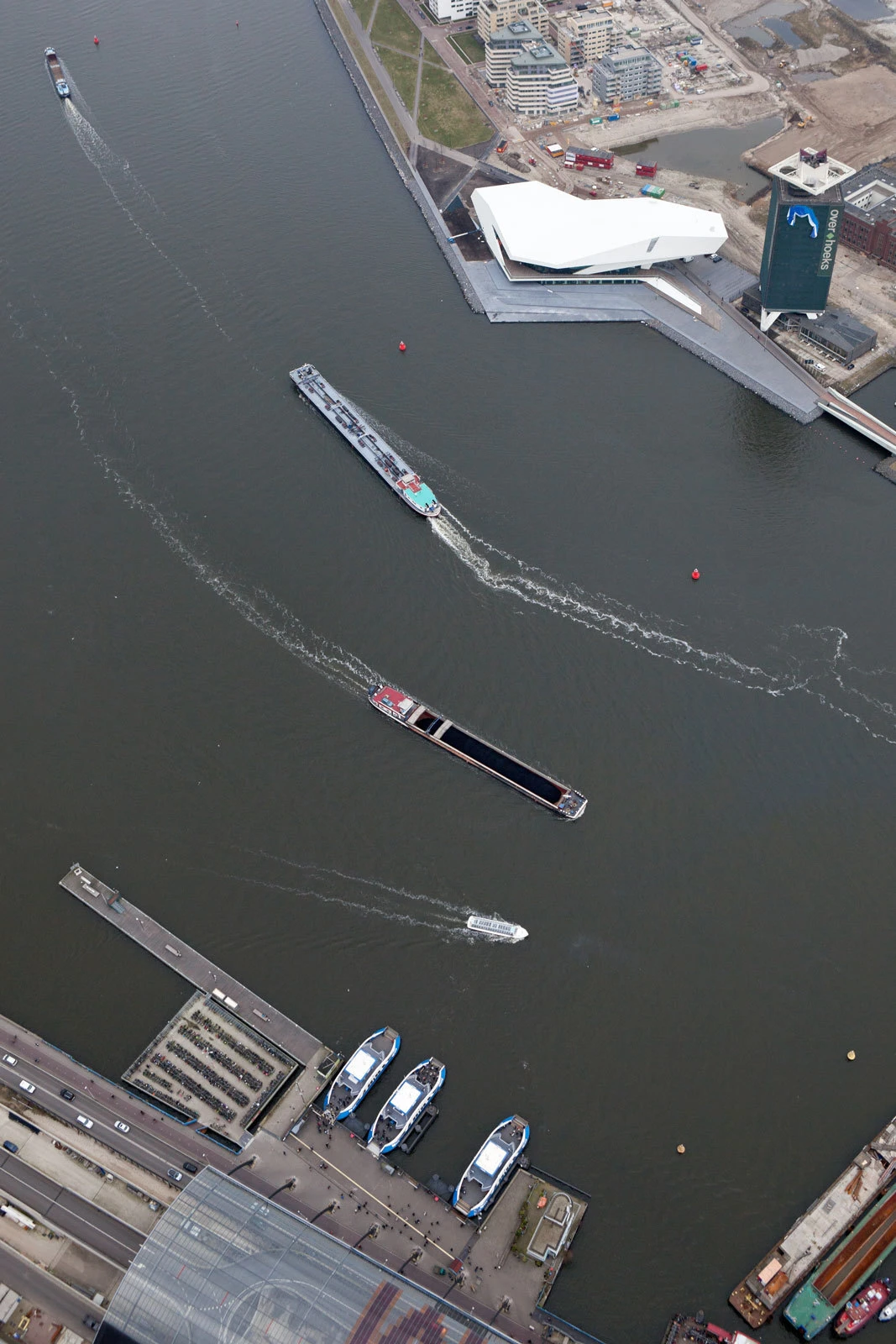 Aerial harbor scene with several motorboats and a long dark barge, beside a white angular waterside building along the quay.