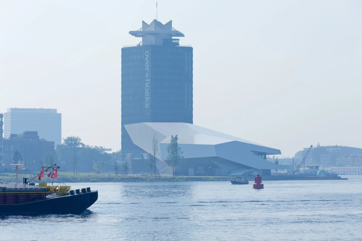 Tall rectangular office tower beside a futuristic angular riverside building; a boat glides on the river.