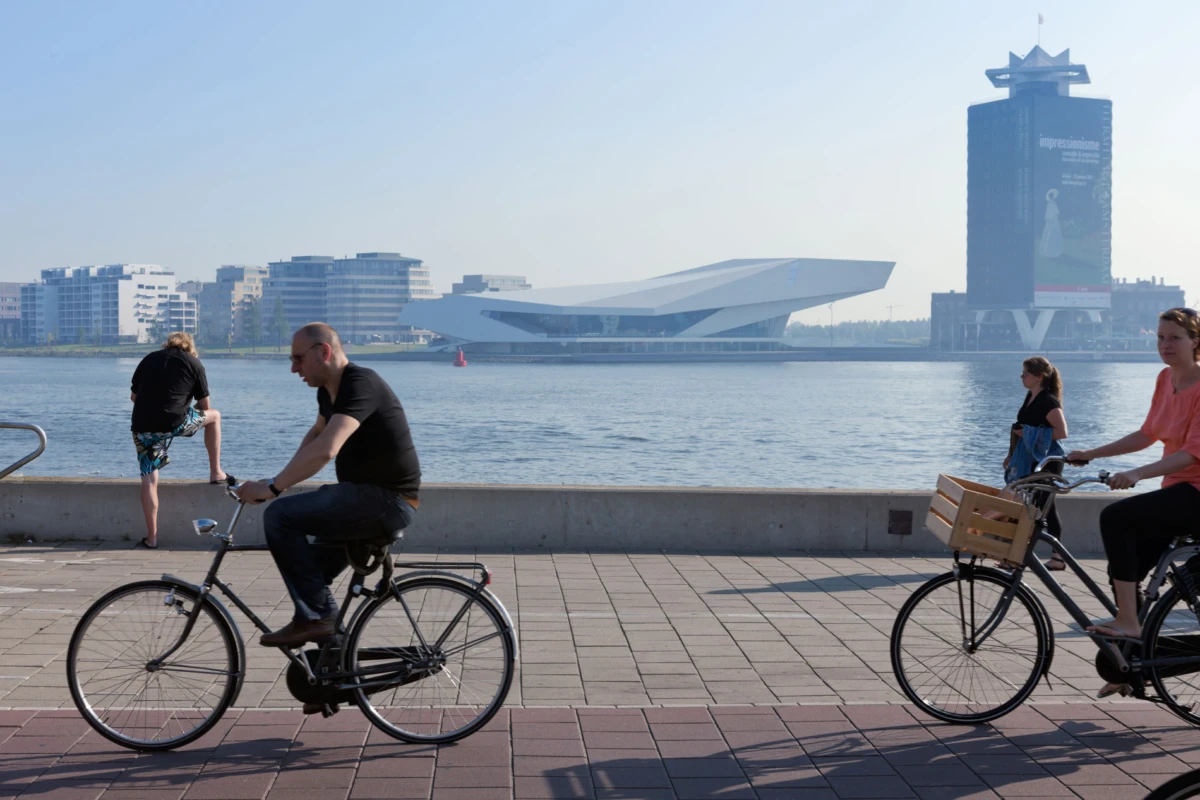 Two cyclists ride along a waterfront promenade by the harbor, with modern buildings across the water.
