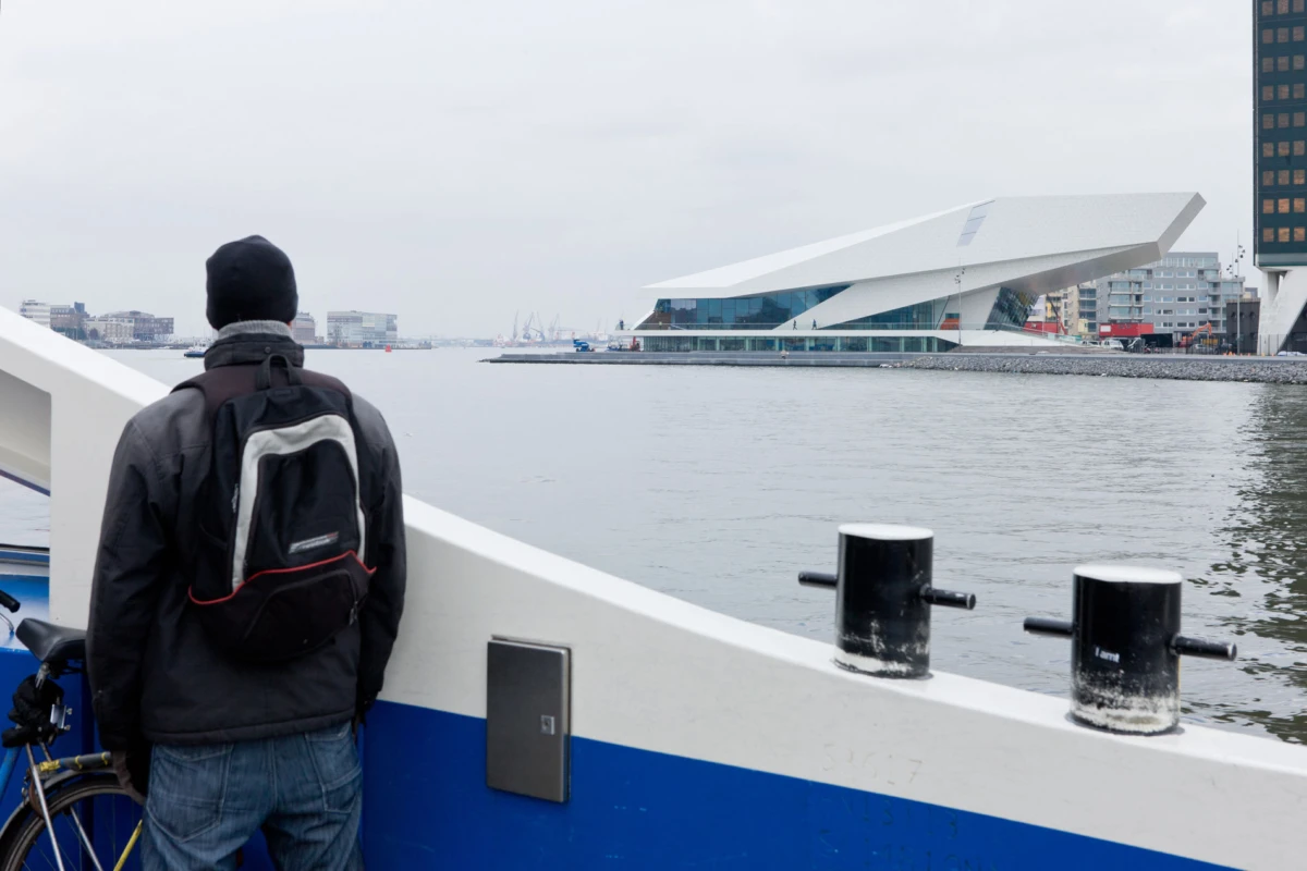 Person wearing a dark jacket and backpack stands at the boat railing by a river, facing a modern angular museum across the water.