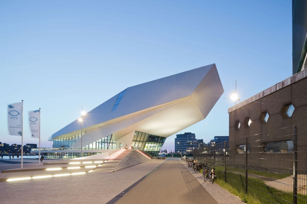 Futuristic white angular museum extends over a waterfront promenade; glass entrance, illuminated stairs, flags, and bicycles along a fence at dusk.