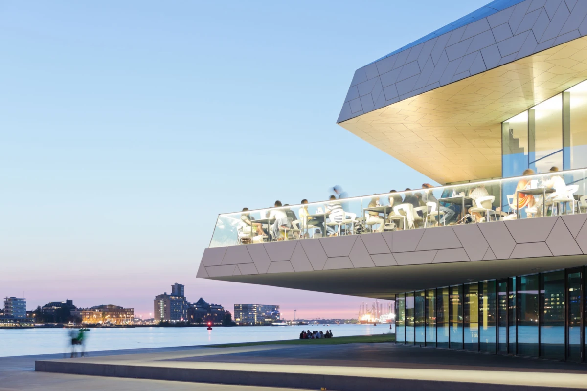 Modern glass-walled restaurant terrace with diners on an outdoor balcony over the water at dusk; city skyline in the distance.