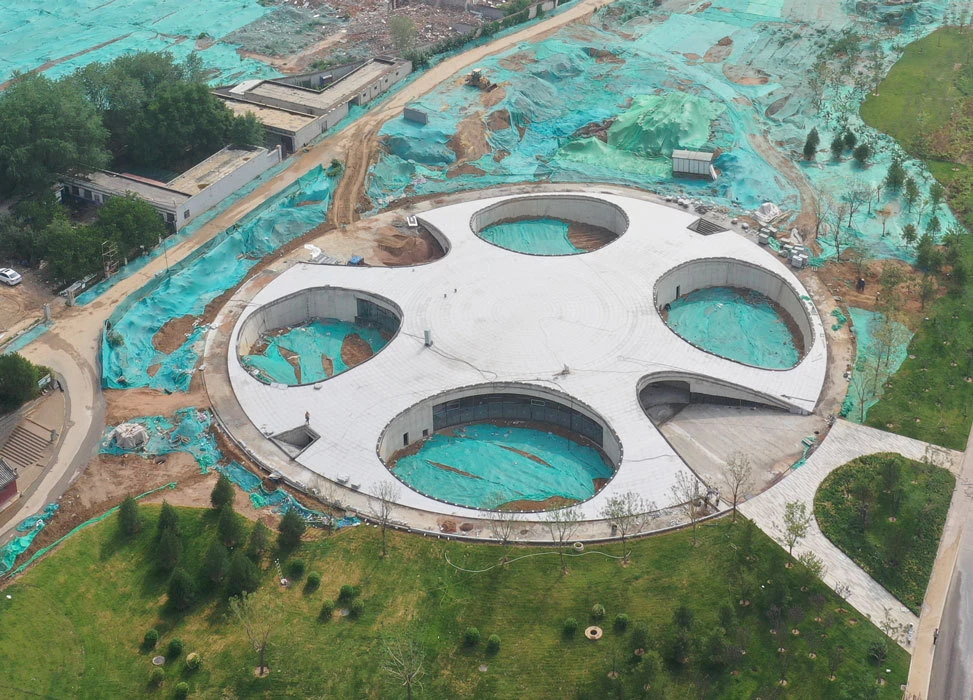 An aerial view of a circular building under construction with four circular courtyards, turquoise site tarps, and surrounding greenery.