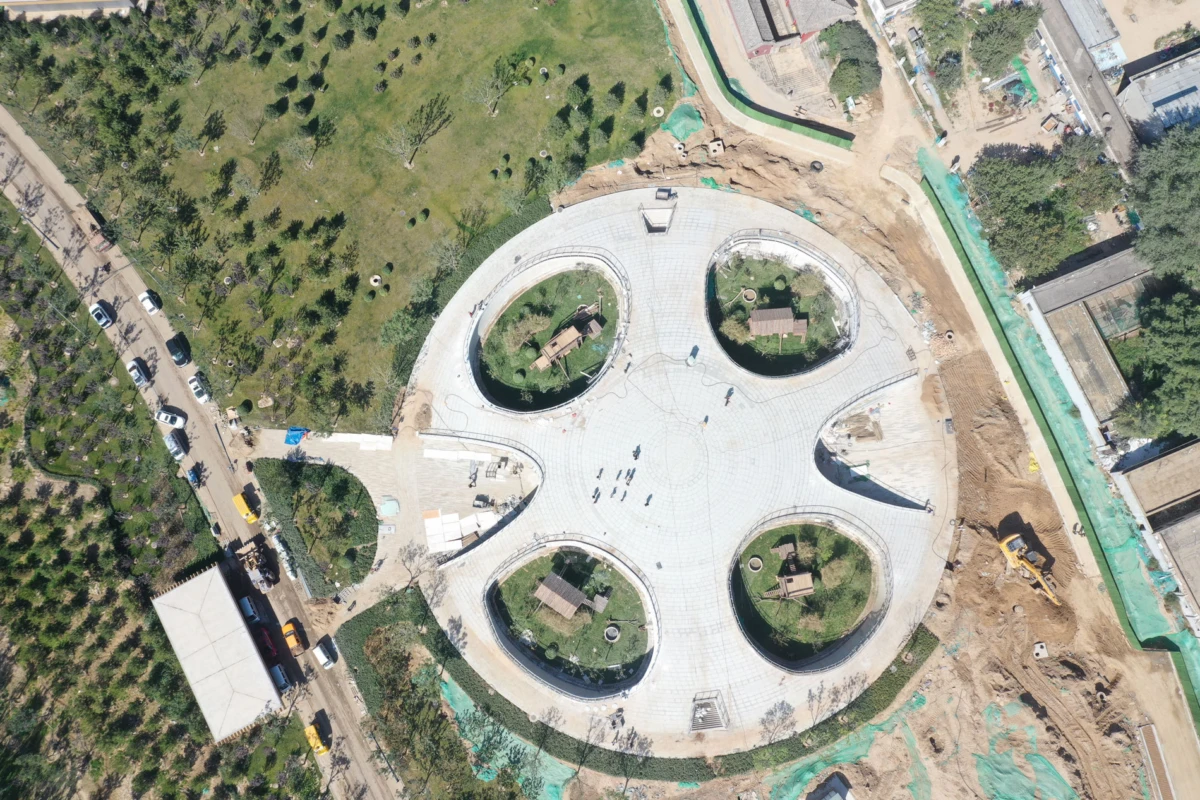 An aerial view of a circular plaza under construction with four circular garden beds; adjacent greenery and roadworks.
