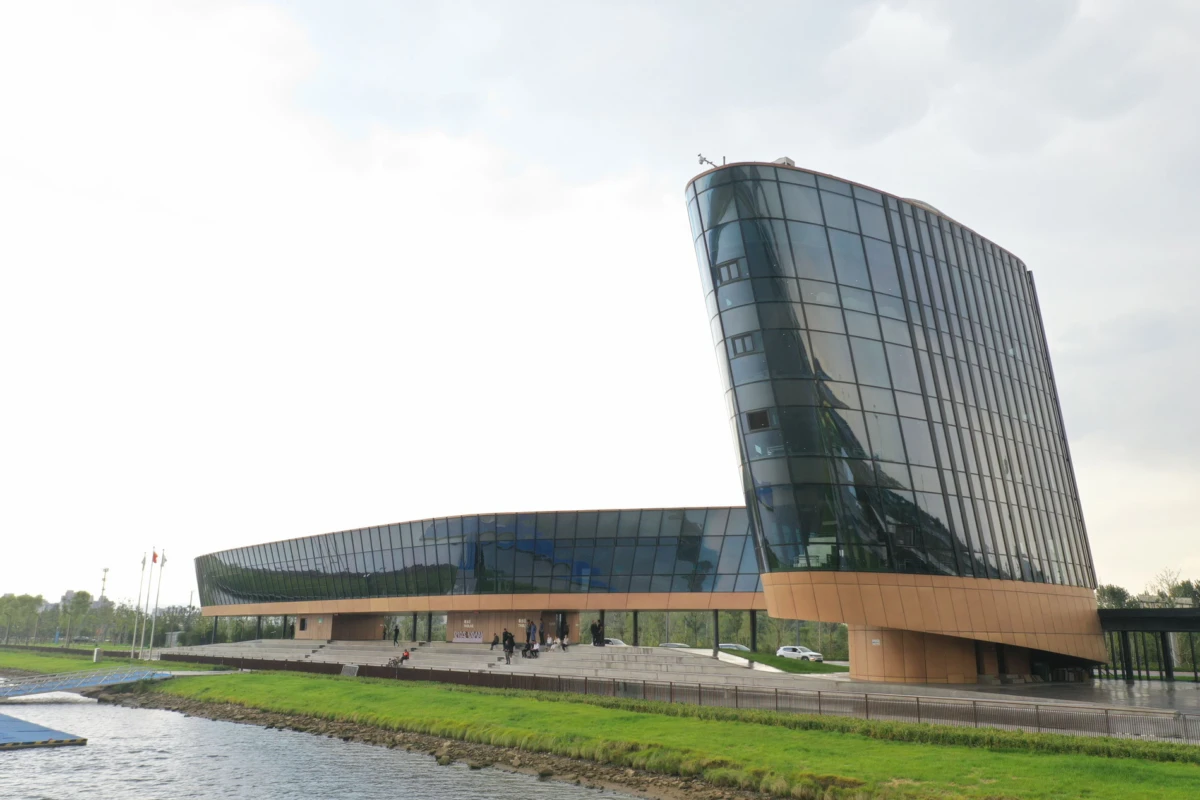 Curved glass office tower beside a river, connected to a low glass annex; pedestrians along the riverside walkway.
