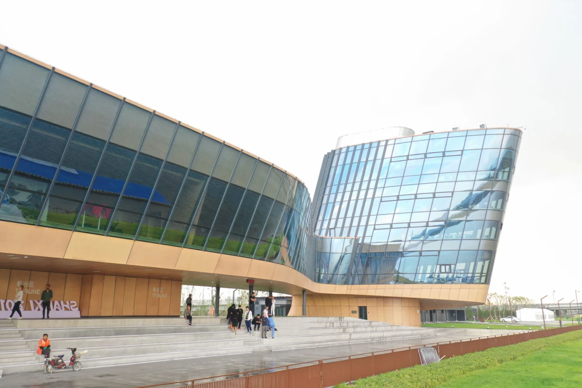 Modern glass-fronted building with a curved facade; people walk up steps to the entrance on a paved plaza with a green lawn nearby.