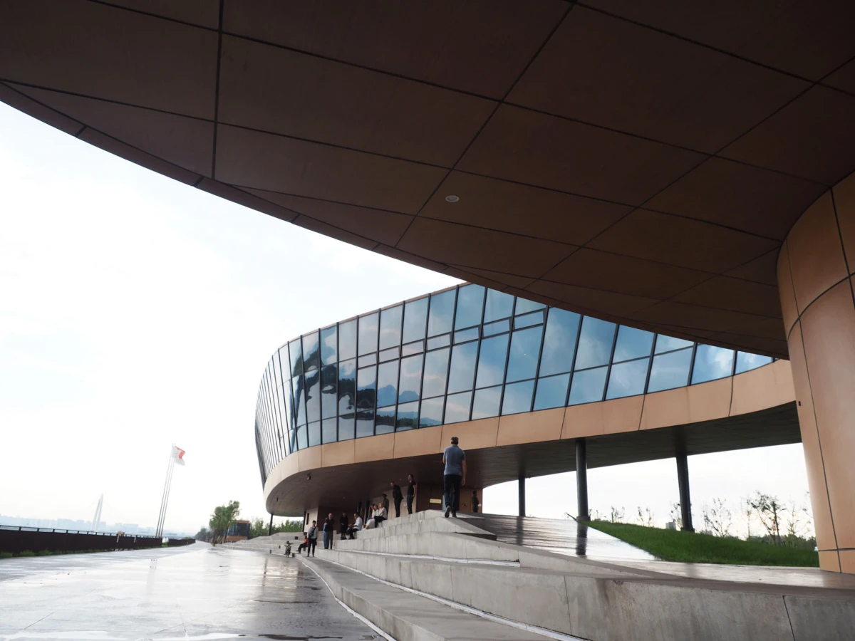 Curved glass-fronted building with people on concrete steps beneath a large overhang in an outdoor plaza.