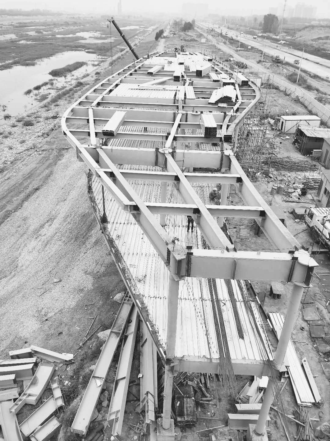 Overhead view of an under-construction steel bridge with scaffolding and a crane, on a muddy construction site along a road.