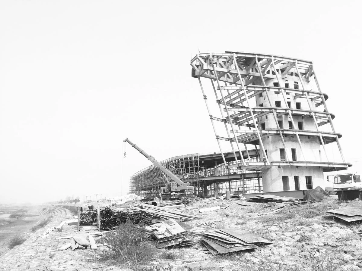 Under-construction multi-story concrete building with external scaffolding; crane on site and piles of lumber on a dirt construction yard.