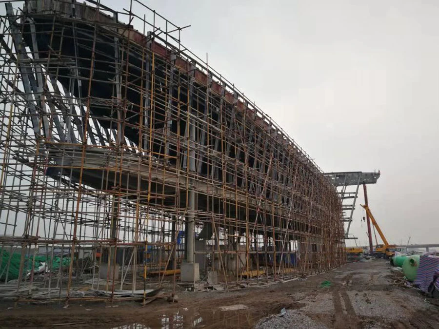 Curved elevated deck under construction, wrapped in bamboo scaffolding at a muddy construction site; crane in the background.