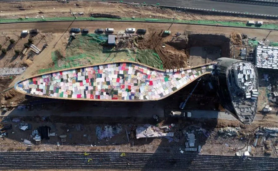 Elongated, hull-shaped building with a multicolored tiled roof at a construction site, surrounded by dirt, machinery, and a nearby road.