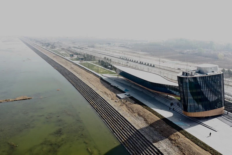 Aerial view of a futuristic waterfront transport hub with a curved white roof and glass-tower building along a long terraced embankment by the river.