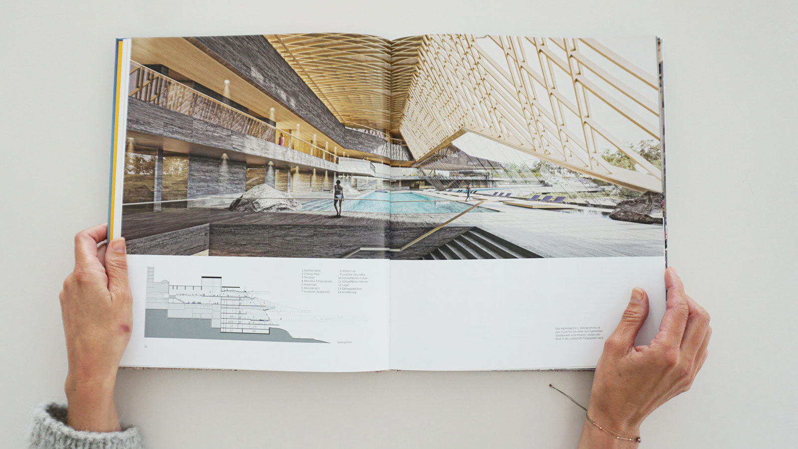 Two hands hold open magazine showing indoor pool architectural rendering in a modern atrium.