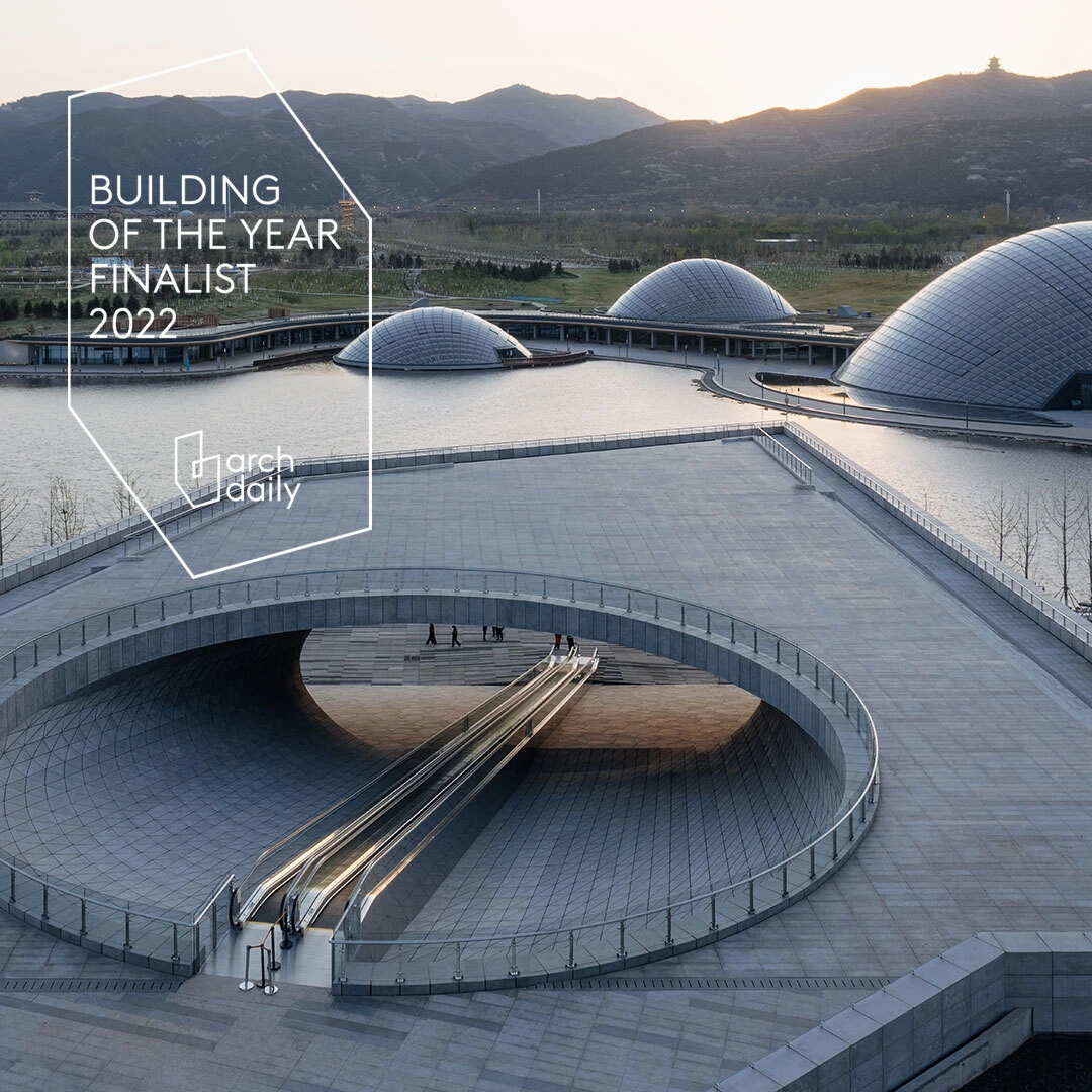 Modern architectural complex with glass domes, a circular ramp, and escalators descending into a tunnel; mountains in the background.
