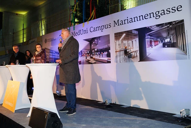 Speaker at podium on stage; panelists at white high-top tables; backdrop reads 'MedUni Campus Mariannengasse' with interior photos.