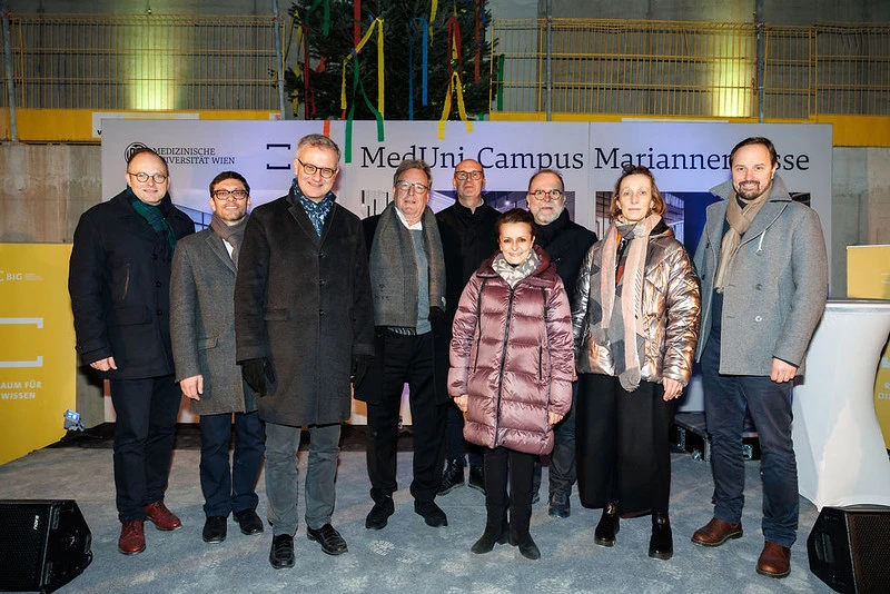 Group of adults in winter coats posing for a photo indoors at a university event, in front of a banner reading MedUni Campus Mariannen.