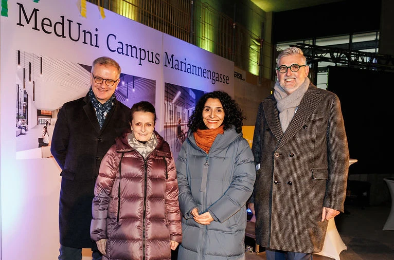 Four adults, two men and two women, posing for a photo in front of a MedUni Campus Mariannengasse banner; wearing winter coats.