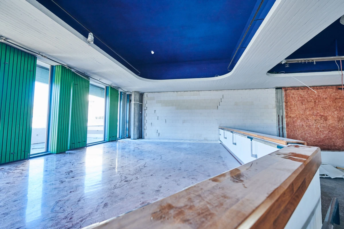 Empty interior room with a curved blue ceiling, white brick walls, green vertical blinds along large windows, and a wooden railing.
