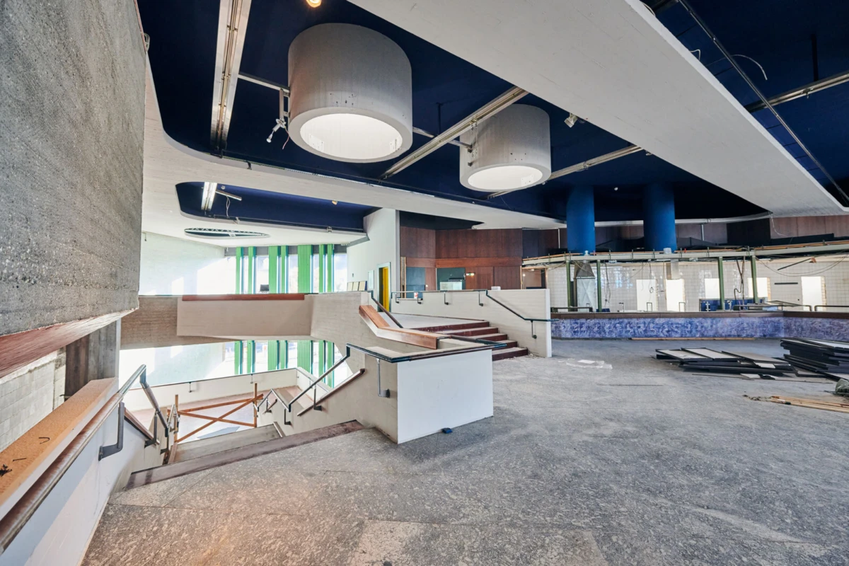 Unfinished multi-level interior lobby under renovation with staircases, ramps, blue columns, and round ceiling lights.