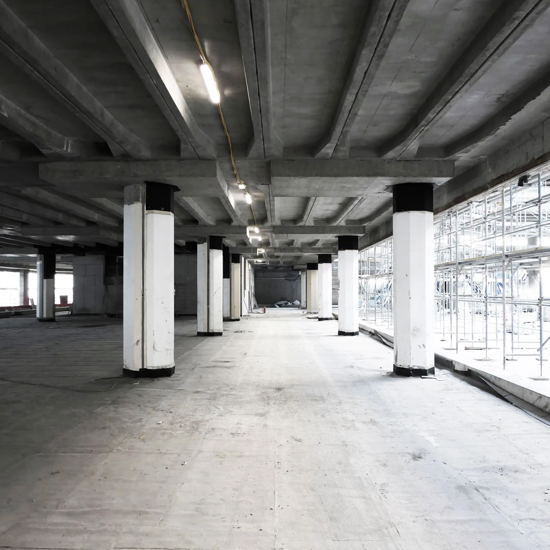 Empty unfinished concrete warehouse interior with rows of white columns and scaffolding along the right side.