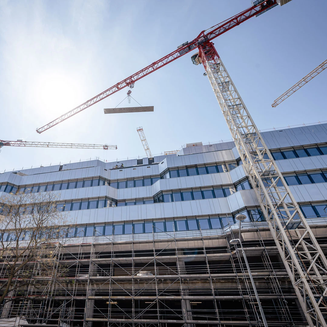 Red-and-white tower crane lifts a concrete slab over a glass-office building under construction, with scaffolding on the lower levels.