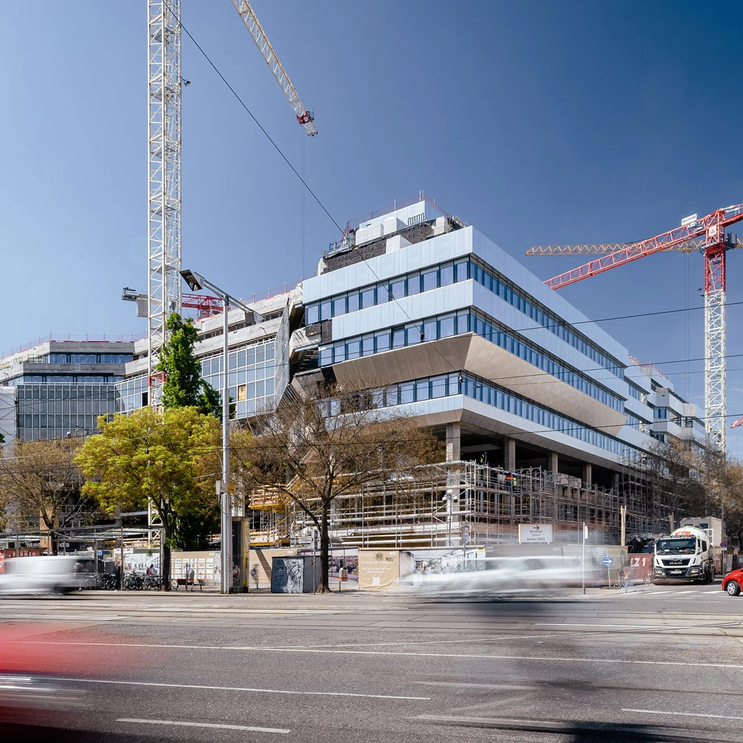 Under-construction glass office building with two cranes over a city street and passing traffic.