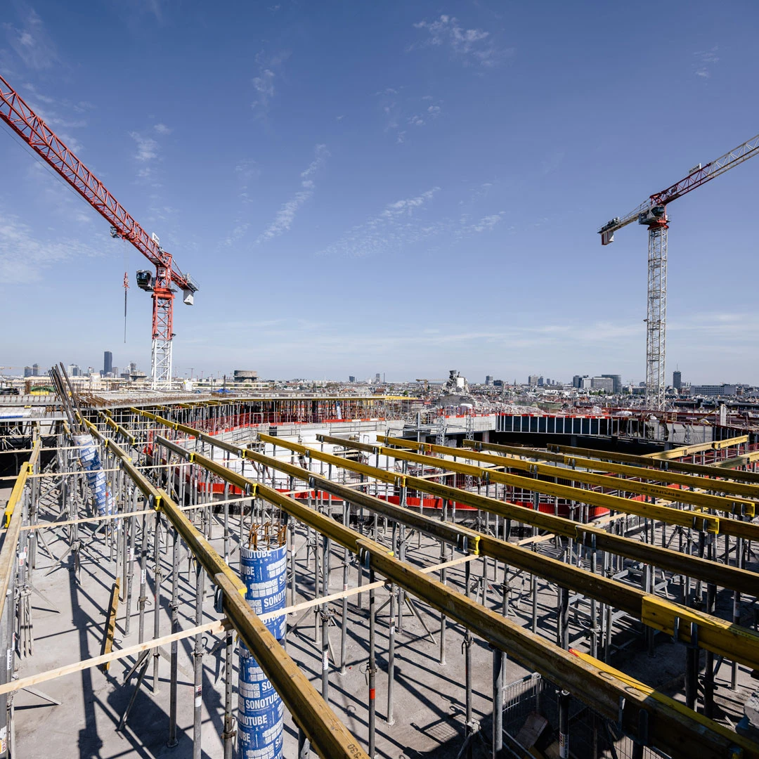Construction site with scaffolding, yellow beams, and two red-and-white tower cranes over an urban skyline.