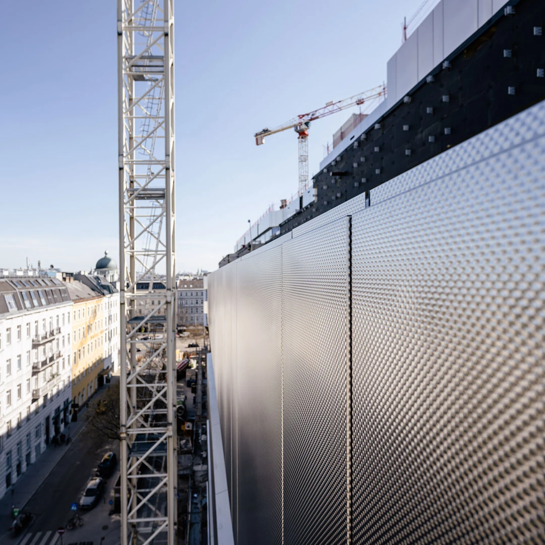 Urban construction site with a tall crane beside a perforated metal wall, overlooking a city street.