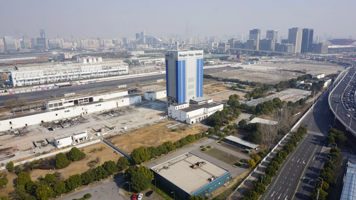 Blue-and-white industrial tower beside a multi-lane highway, with rail yard and urban skyline in the distance.