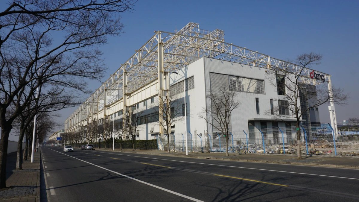 Large industrial warehouse with white facade and steel lattice framework on the roof under construction beside a tree-lined street.