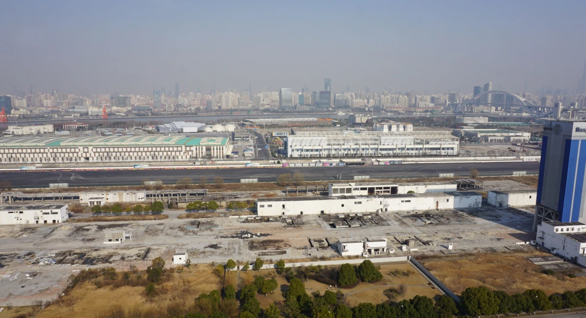 Aerial view of an industrial complex with white warehouses, vacant lots, and a tall blue building on the right, with a distant city skyline.