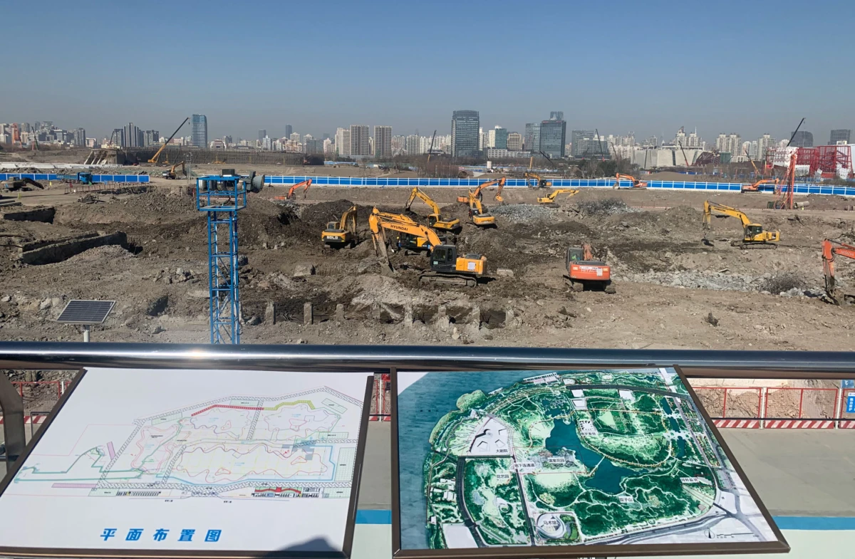 Construction site with multiple excavators digging soil; foreground shows plan boards; city skyline in the background.