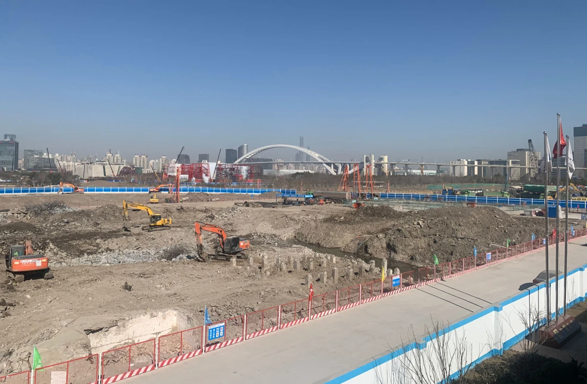 Construction site with excavators digging in dirt; urban skyline with an arched bridge in the distance.