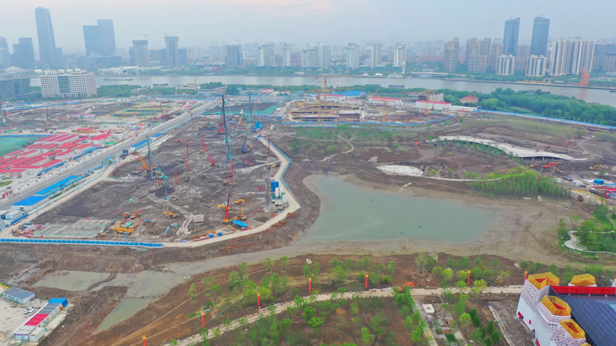 Aerial view of a large construction site with cranes and excavators at work, muddy terrain, a winding river, and a skyline of high-rise buildings.