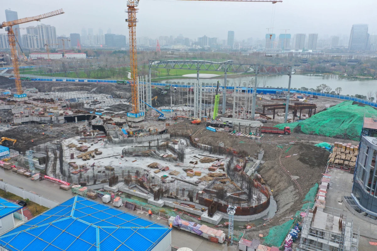 Construction site with two tall cranes over a circular foundation under construction, with urban skyline and river in the background.