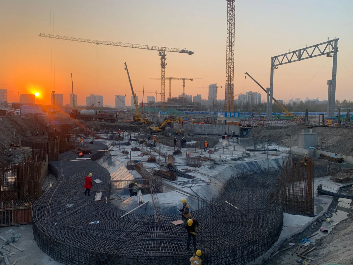 Construction site at sunset with workers assembling a circular reinforced concrete foundation; cranes loom over the site.
