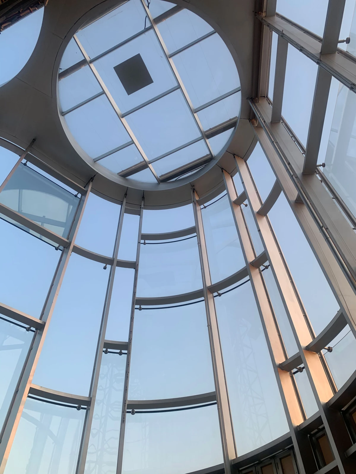 Interior of circular glass atrium, seen from below, with steel frames and a square skylight overhead.