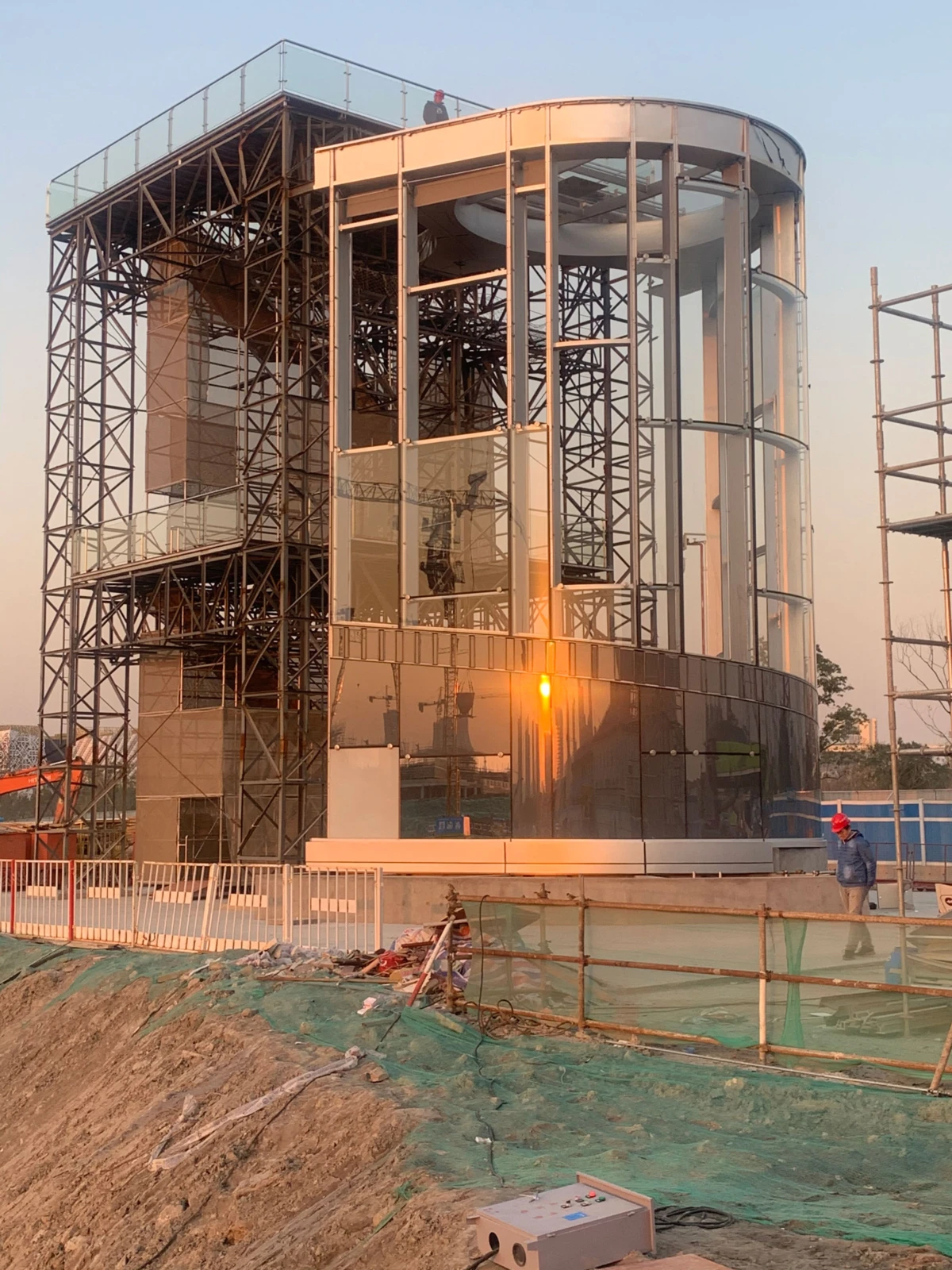 Glass-clad cylindrical building under construction with steel scaffolding; worker in a red helmet stands on the top platform at sunset.