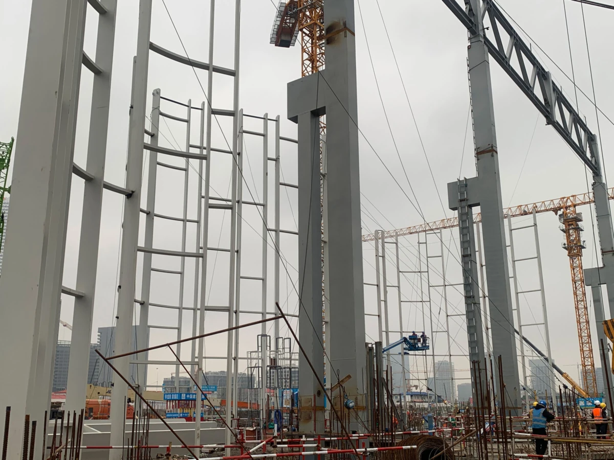 Construction site with tall steel frame columns, scaffolding, and cranes; workers on an elevated platform assembling the structure.
