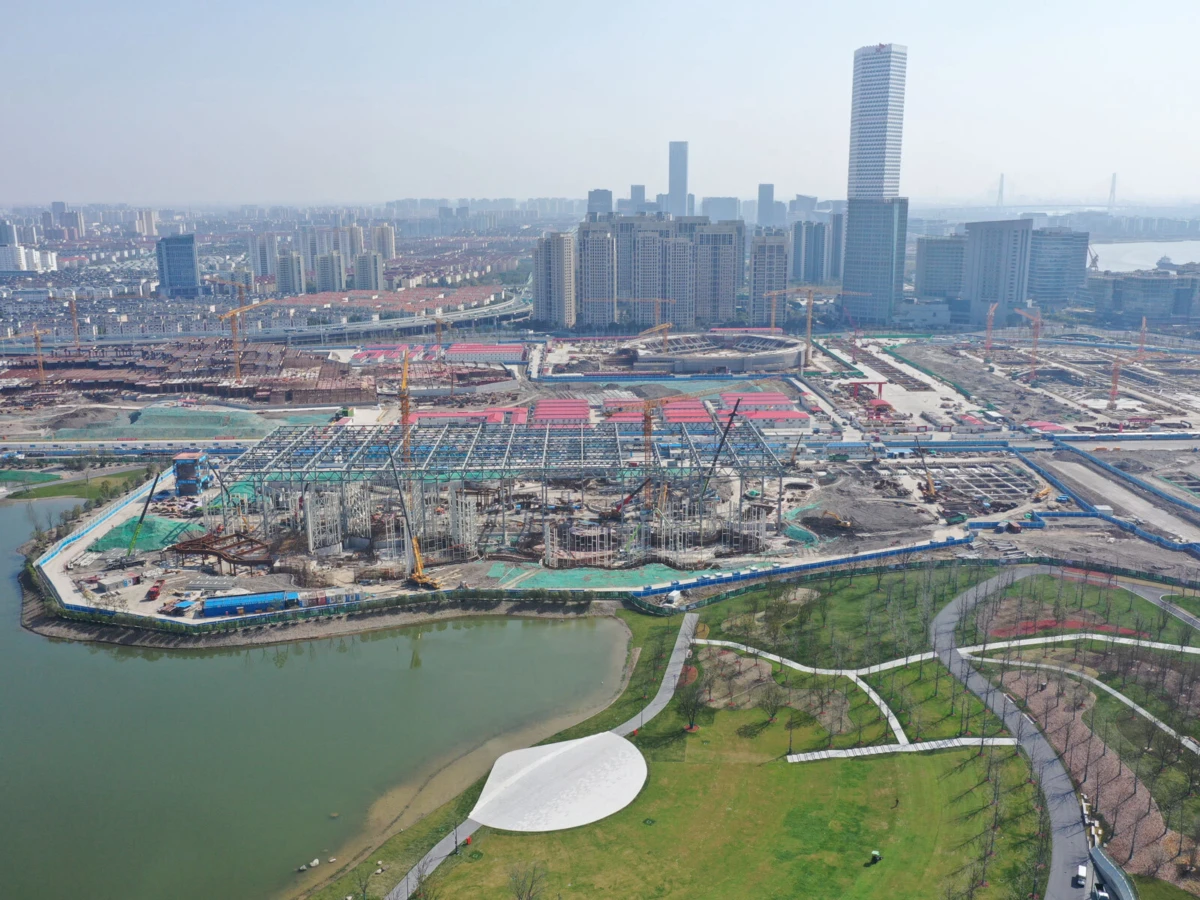 Construction site with cranes and steel framework along a river; city skyline in the background and a curved park with a white pavilion in the foreground.