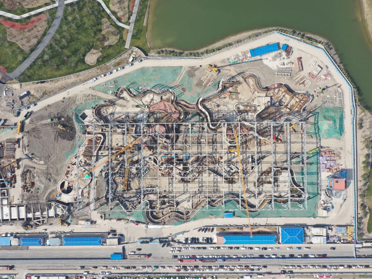 Aerial view of a roller coaster under construction inside a fenced site by a body of water, with cranes and tangled steel tracks.