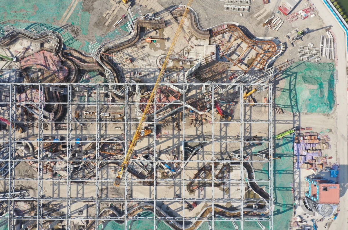 Aerial view of a construction site with a steel grid frame, extensive twisting pipelines, and a yellow crane spanning the area.